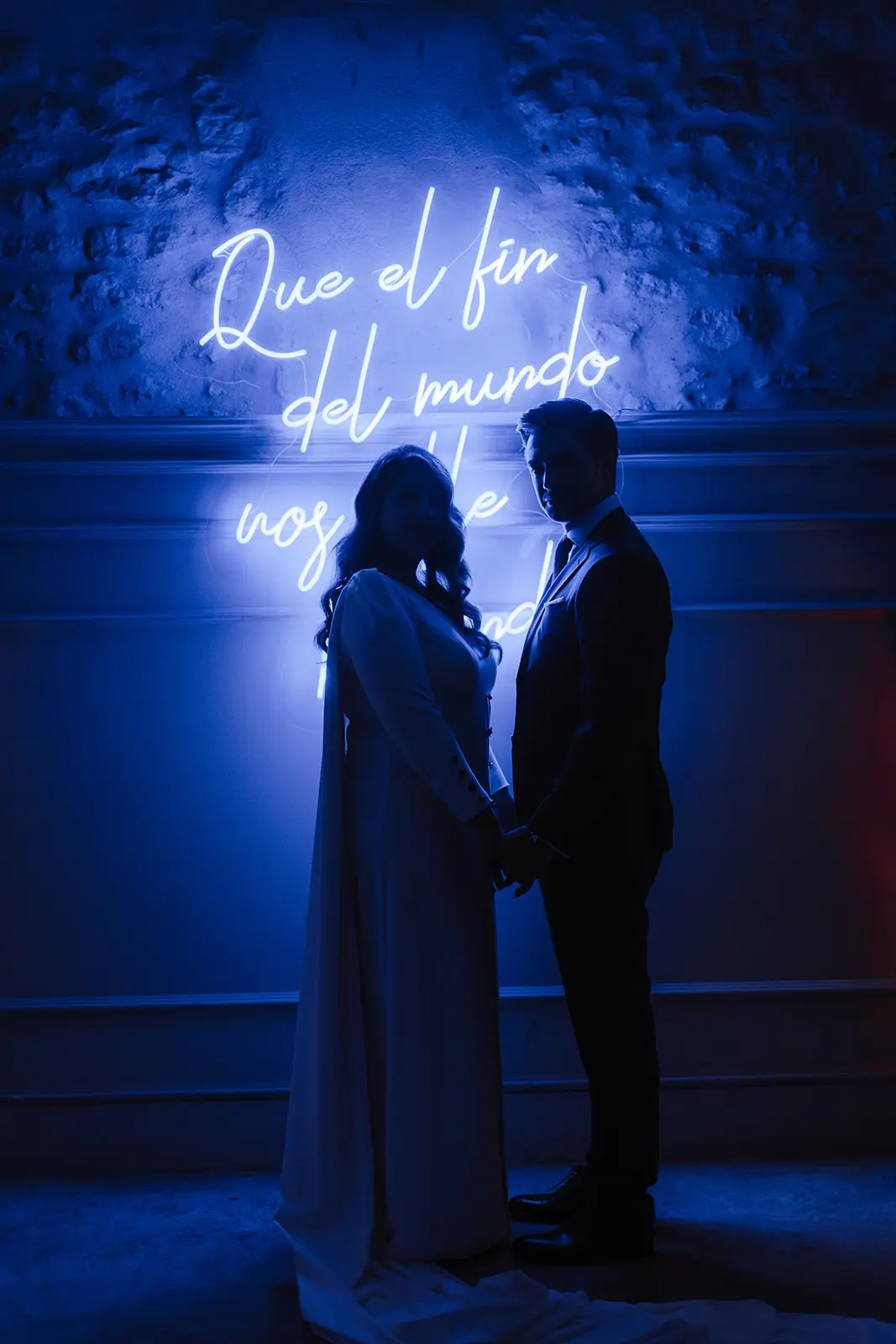 Pareja de novios mirando a la cámara bajo el resplandor del neón azul con frase romántica.