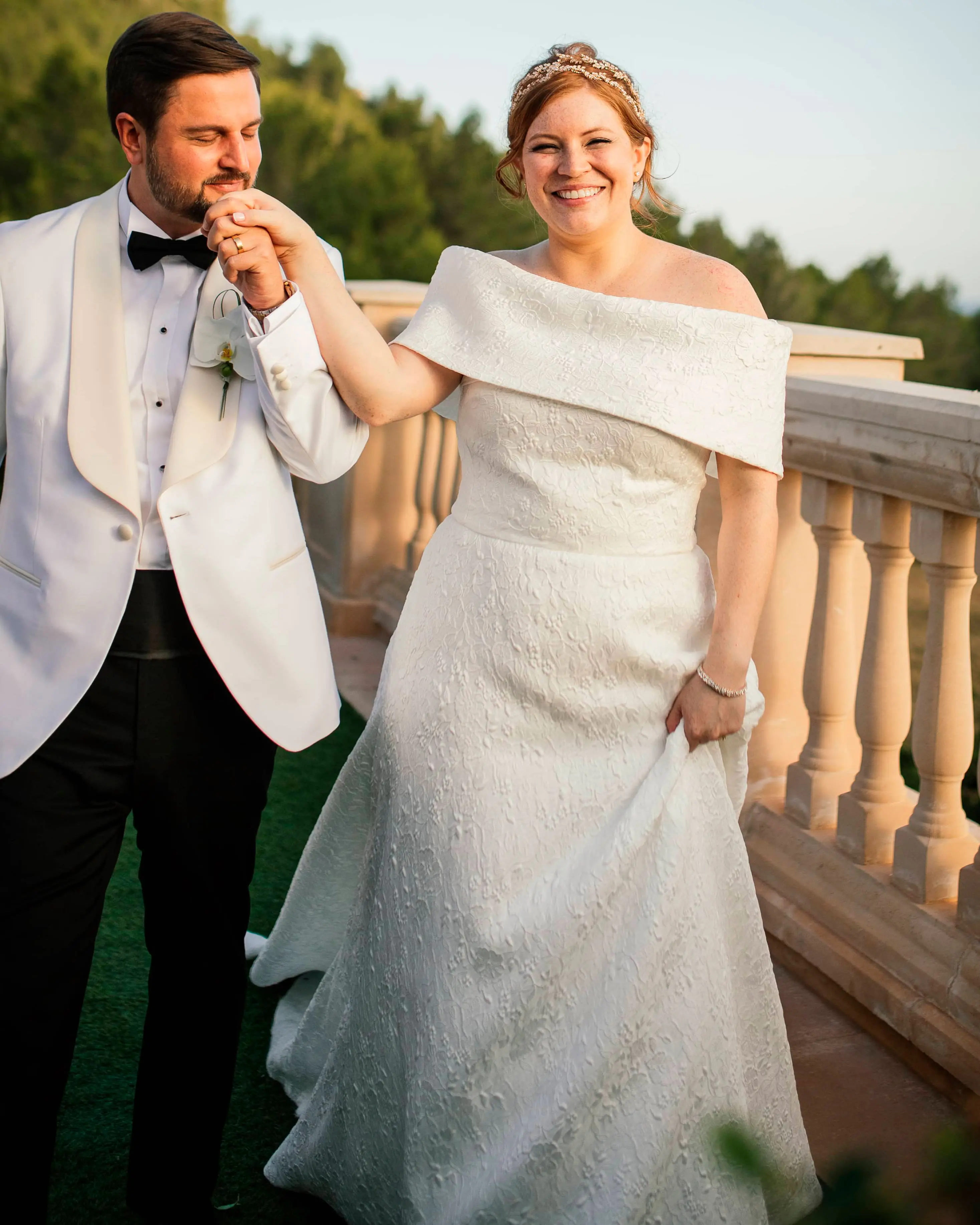 Mau kisses Stephanie's hand, both dressed in wedding attire, in a romantic scene with the beautiful natural landscape of Mallorca in the background.