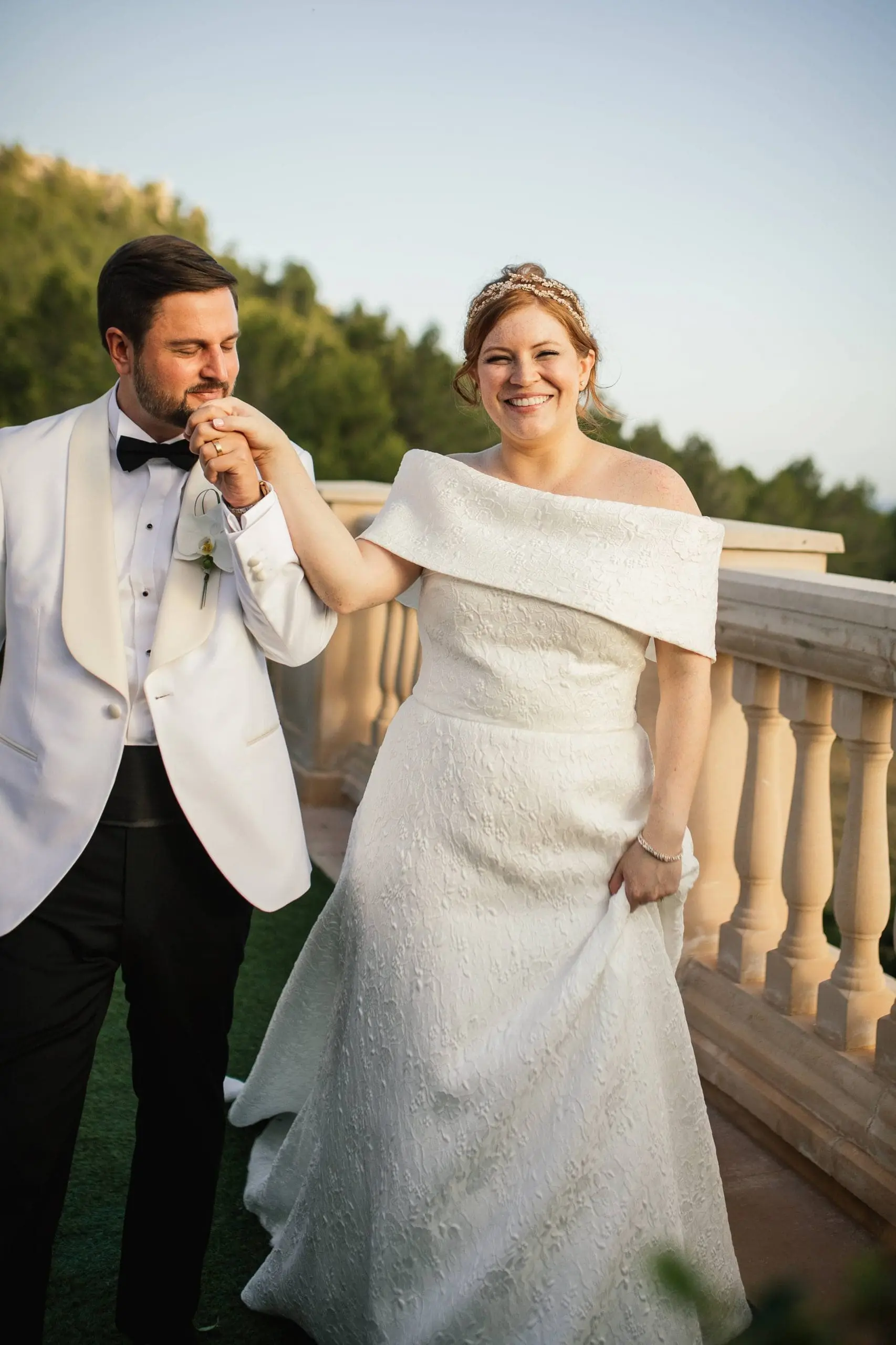 Mau besa la mano de Stephanie, ambos vestidos de novios, con el paisaje natural de Mallorca de fondo.