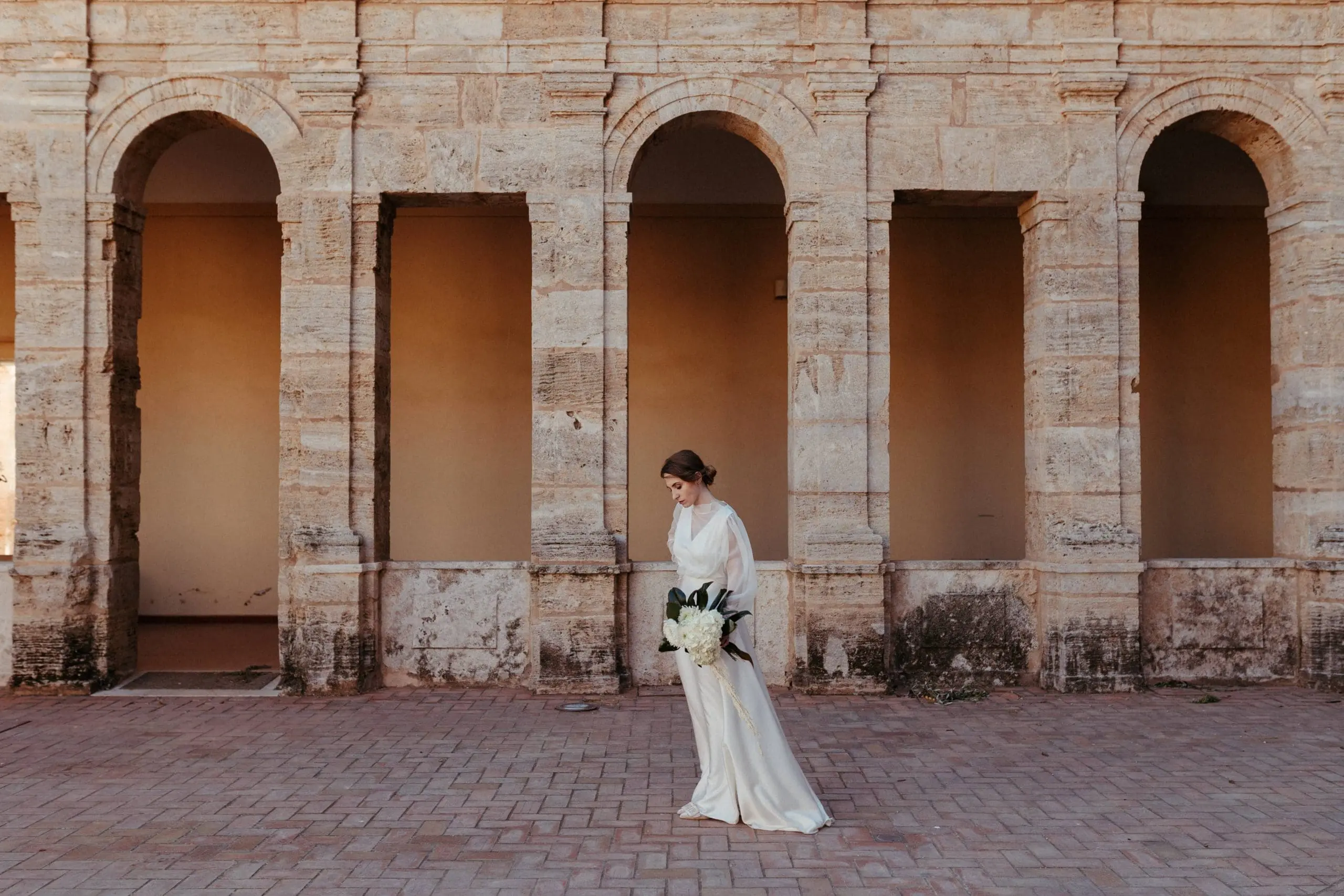 Novia sosteniendo un ramo blanco, frente a los imponentes arcos de la Cartuja de Ara Christi.