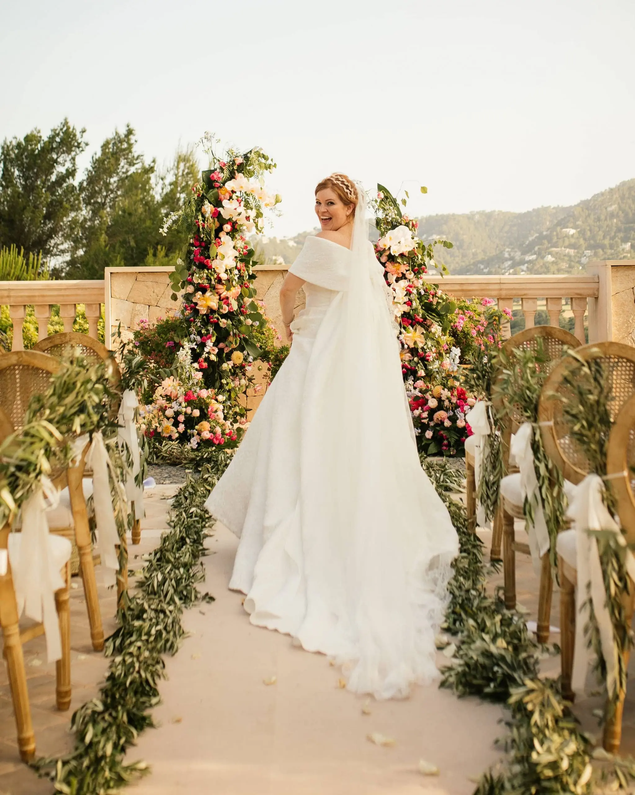 Novia avanzando hacia el altar con semiarcos florales y un sendero de ramas de olivo, con la sierra de Mallorca de fondo.