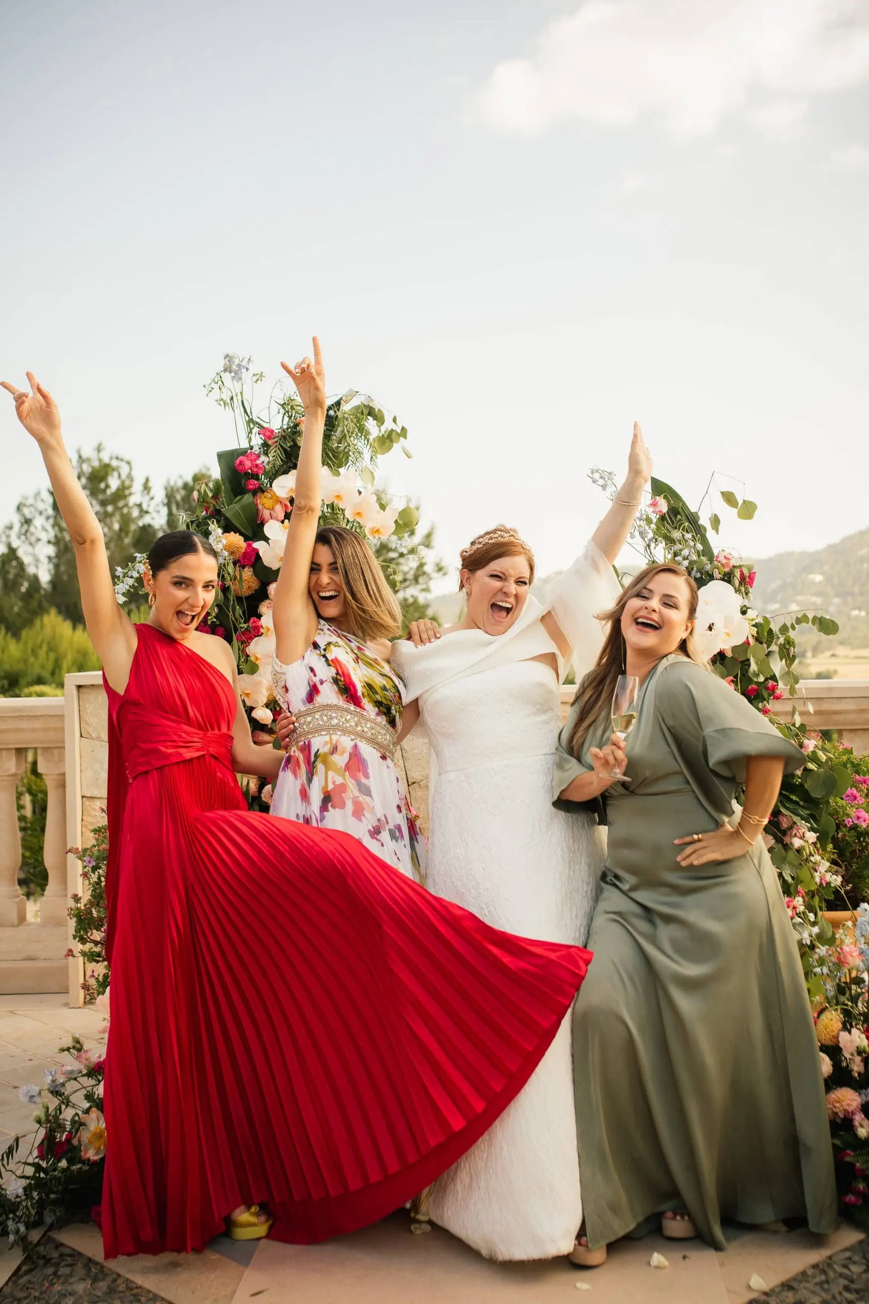 Novia y sus amigas levantan las manos entre dos semiarcos de flores de su ceremonia en Mallorca
