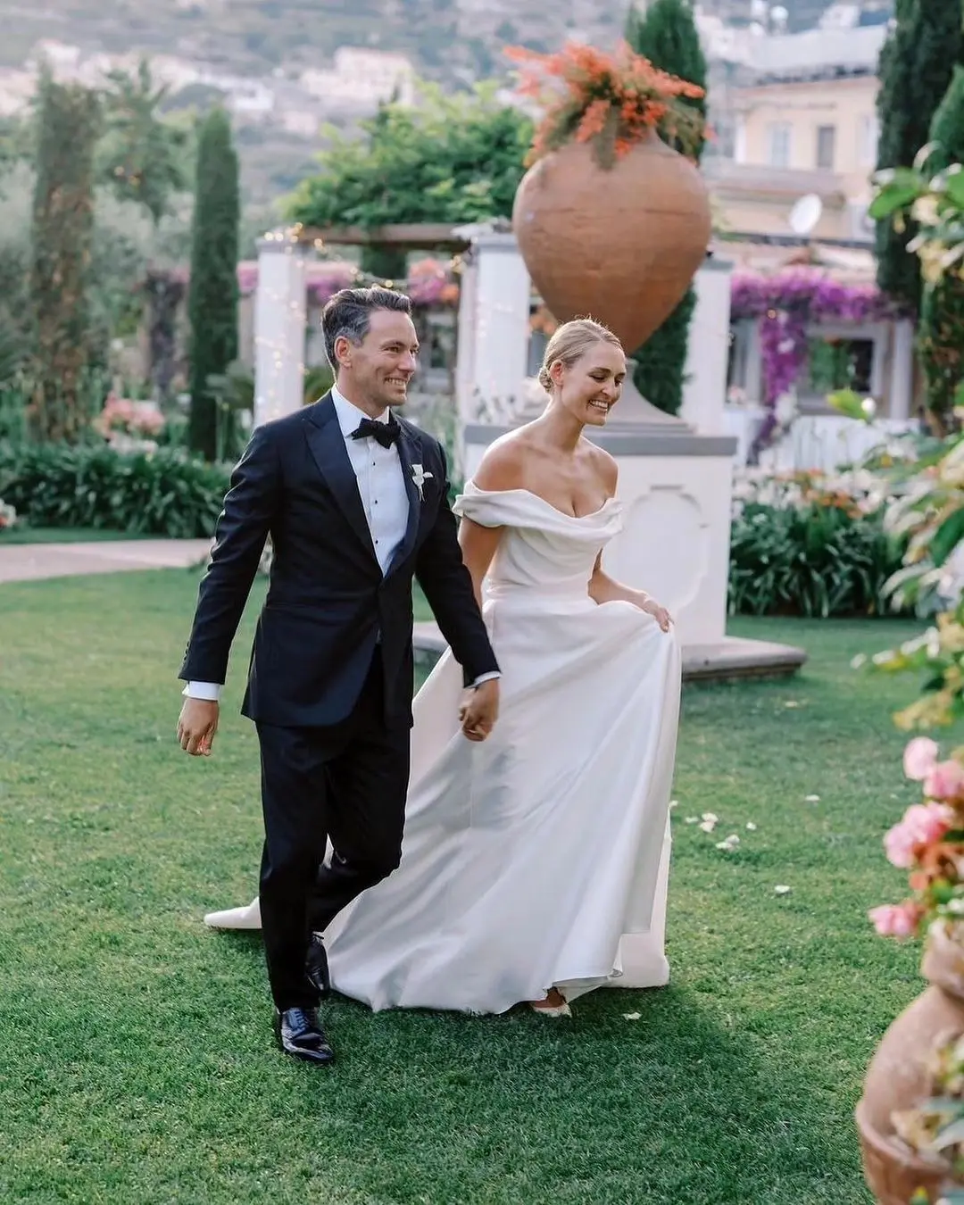 Pareja de novios caminando de la mano por los jardines de Villa Eva en la Costa Amalfitana, con la elegante arquitectura de la villa y el mar brillante al fondo.