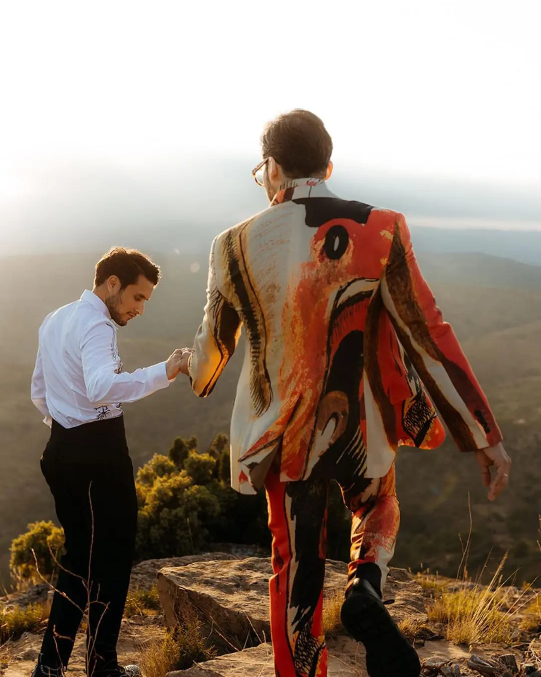 Pareja de chicos en una aventura romántica por un barranco, uno con traje llamativo, celebrando el amor y la libertad en San Valentín.