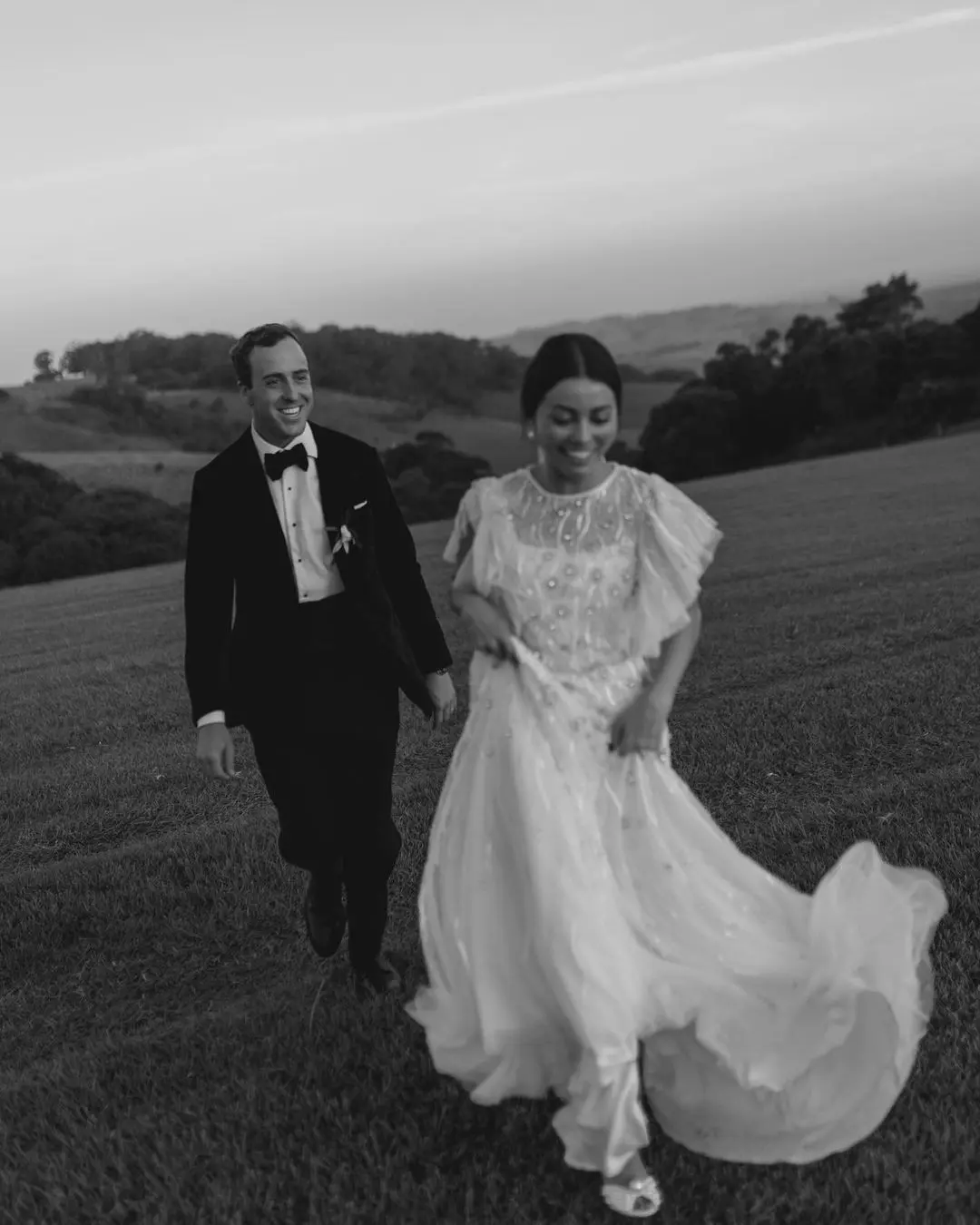 Novios sonriendo y caminando de la mano por un prado, reflejando cómo la tecnología en la organización de bodas enriquece la conexión entre las parejas.