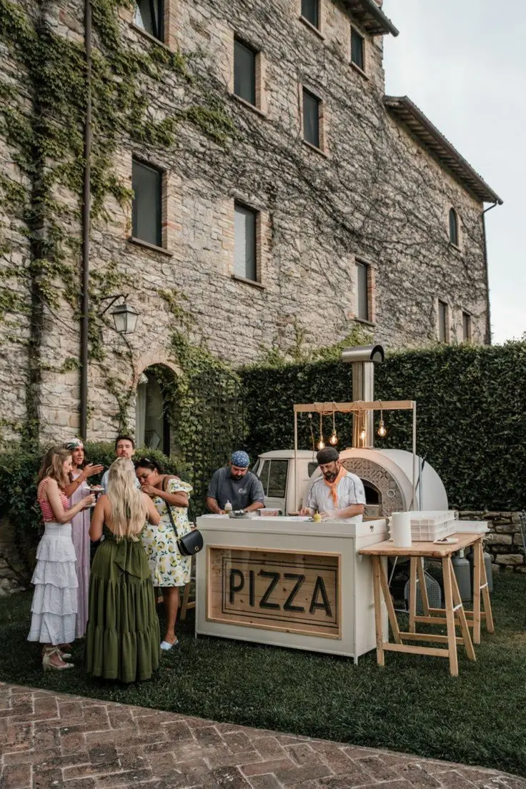 Puesto de pizza al aire libre con invitados disfrutando, opción perfecta para una recena de boda informal.
