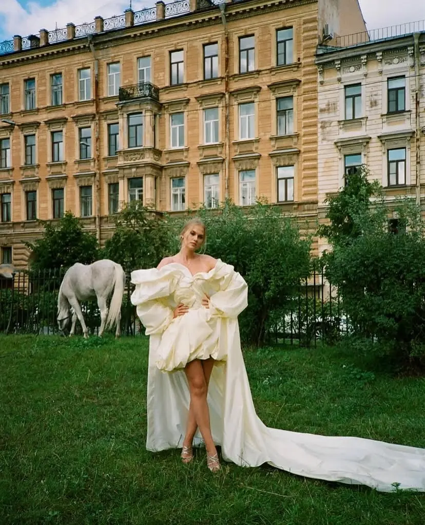 Novia en un jardín con un vestido de diseño único, acompañado por un caballo blanco y un fondo arquitectónico clásico.