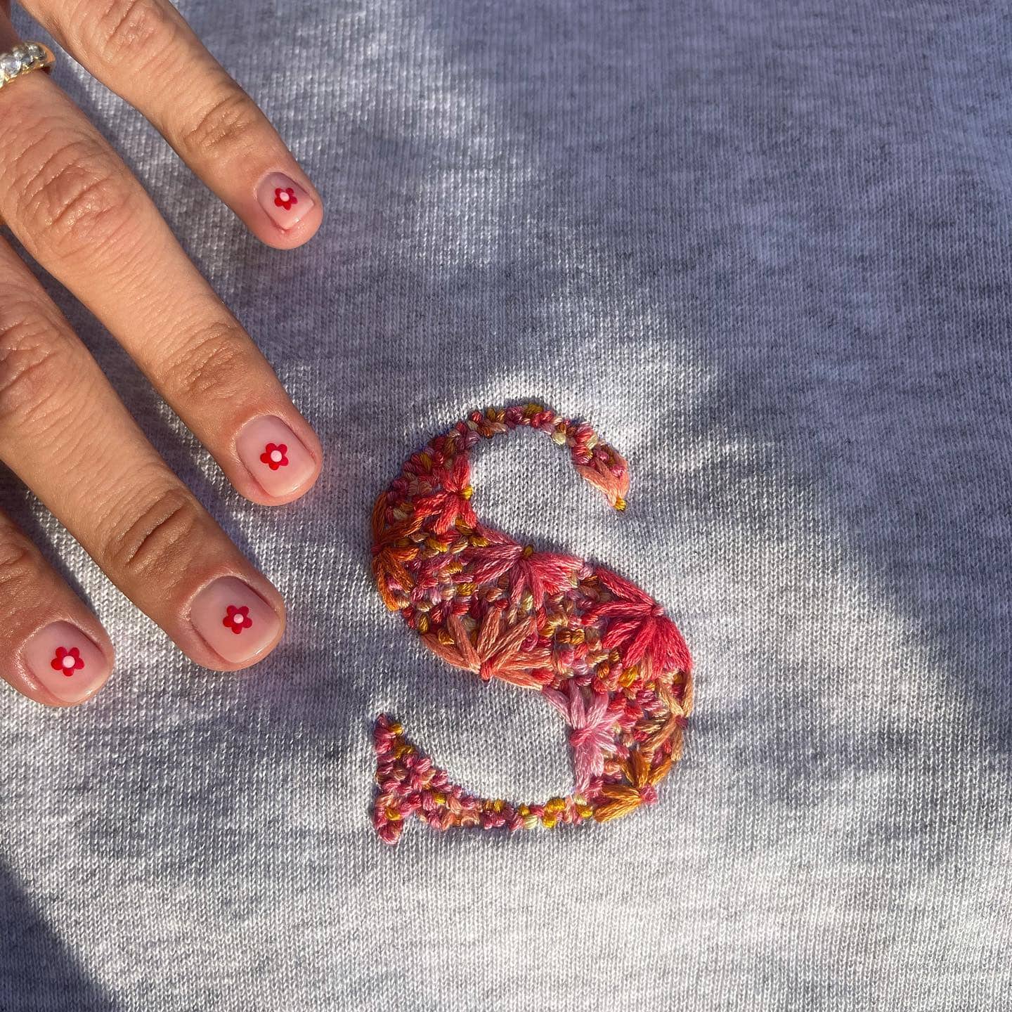 Regalos para invitados: Sudadera gris con bordado personalizado en tonos rojos de la letra 'S' mayúscula. También se aprecian uñas con margaritas pintadas.