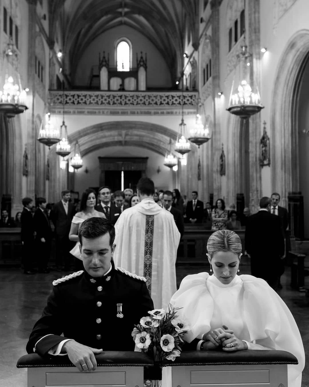 Novios arrodillados rezando en una iglesia durante el rito de la velación, mientras el cura habla a los invitados - Bouclé Weddings
