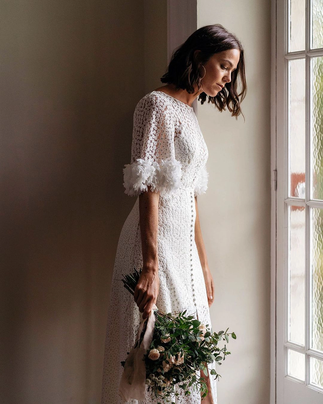 Novia sosteniendo un ramo de flores mientras mira pensativamente por la ventana, reflexionando sobre sus votos matrimoniales.