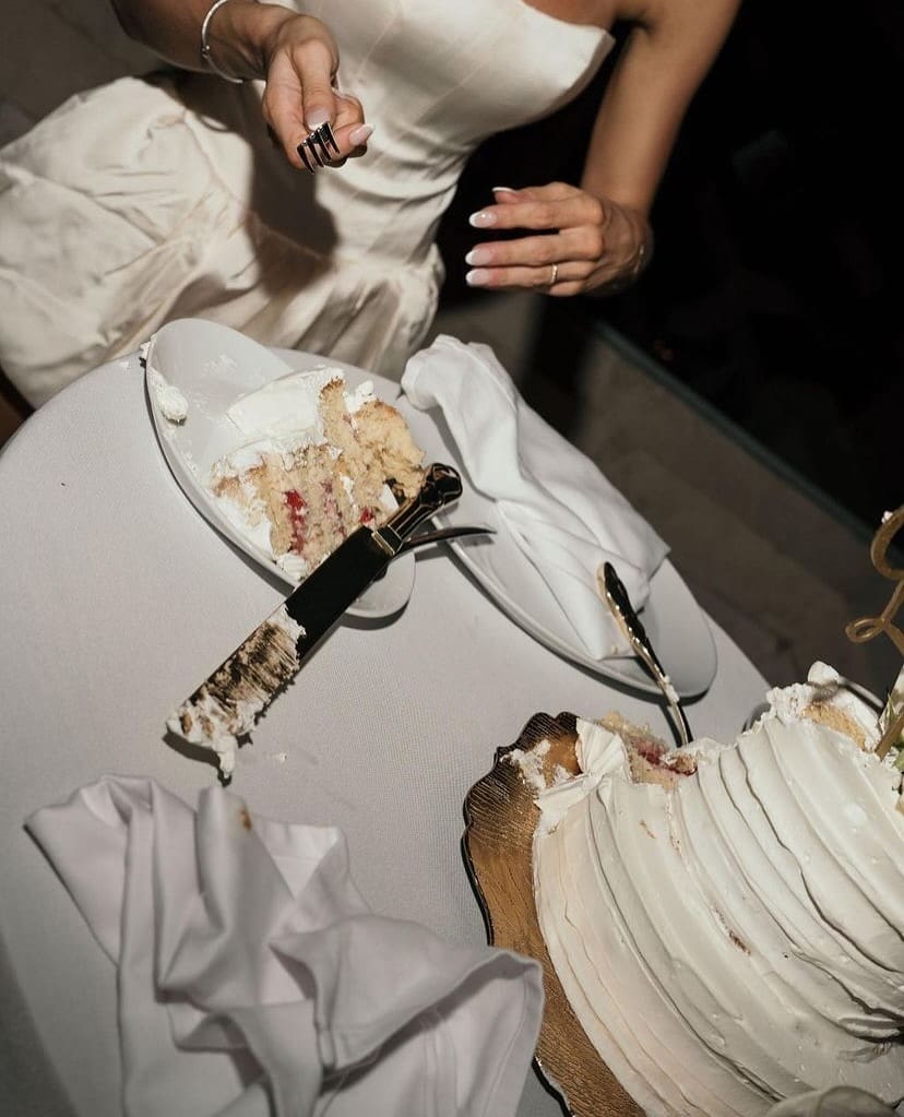 Porción de pastel de bodas servido en un plato mientras una novia sostiene un cuchillo, destacando la celebración del matrimonio.