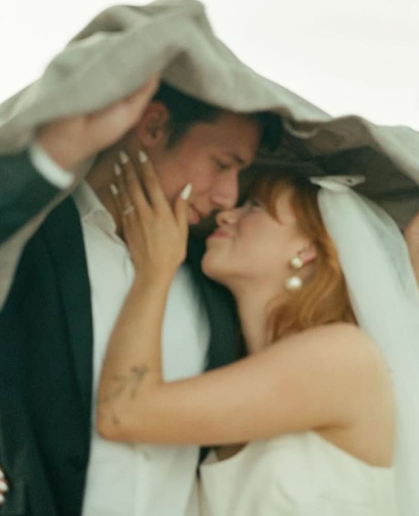 Pareja de recién casados refugiándose bajo una chaqueta con mirada cómplice