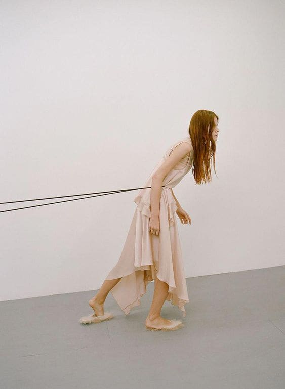 Mujer en vestido claro caminando mientras algo invisible la retiene por la espalda.